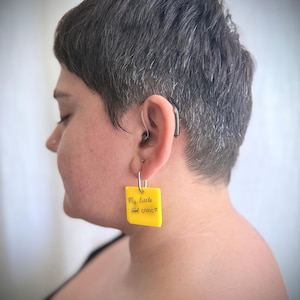 Op de afbeelding: Een vierkante gele acryl oorbel met een zilverkleurige metalen ring. De oorbel toont de zwarte handgeschreven tekst "My little food critic" gevolgd door een klein hartsymbool, hangend aan een zilverkleurige metalen oorbelhaakje.