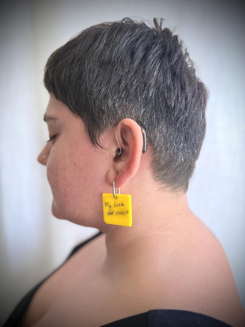 Op de afbeelding: Een vierkante gele acryl oorbel met een zilverkleurige metalen ring. De oorbel toont de zwarte handgeschreven tekst "My little food critic" gevolgd door een klein hartsymbool, hangend aan een zilverkleurige metalen oorbelhaakje.