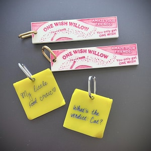 Op de afbeelding: Twee rechthoekige witte sleutelhangers met roze tekst ONE WISH WILLOW, AMAZE YOUR FRIENDS, en You only get ONE WISH, plus twee vierkante gele sleutelhangers met handgeschreven zwarte tekst My little food critic en What's the verdict: Cat?