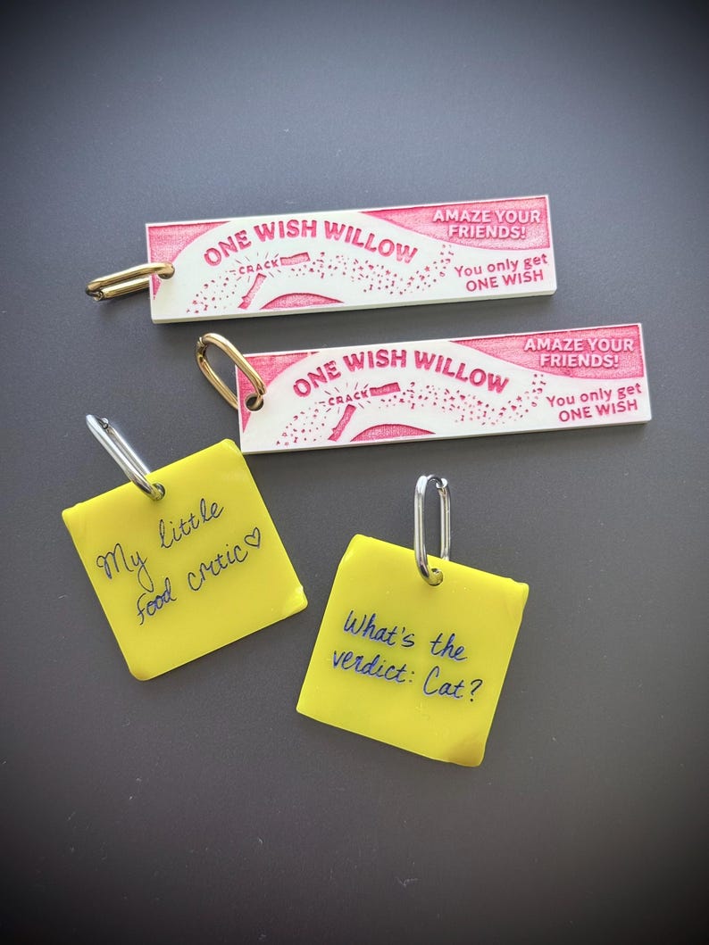 Op de afbeelding: Twee rechthoekige witte sleutelhangers met roze tekst ONE WISH WILLOW, AMAZE YOUR FRIENDS, en You only get ONE WISH, plus twee vierkante gele sleutelhangers met handgeschreven zwarte tekst My little food critic en What's the verdict: Cat?