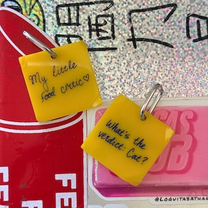 Op de afbeelding: Twee vierkante gele bedels met zilverkleurige metalen ringetjes. E&eacute;n bedel bevat de zwarte handgeschreven tekst "My little food critic♡" en de andere bedel bevat de zwarte handgeschreven tekst "What's the verdict: Cat?" in een cursief lettertype.