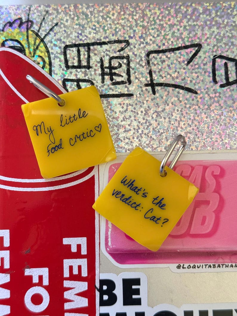 Op de afbeelding: Twee vierkante gele bedels met zilverkleurige metalen ringetjes. E&eacute;n bedel bevat de zwarte handgeschreven tekst "My little food critic♡" en de andere bedel bevat de zwarte handgeschreven tekst "What's the verdict: Cat?" in een cursief lettertype.