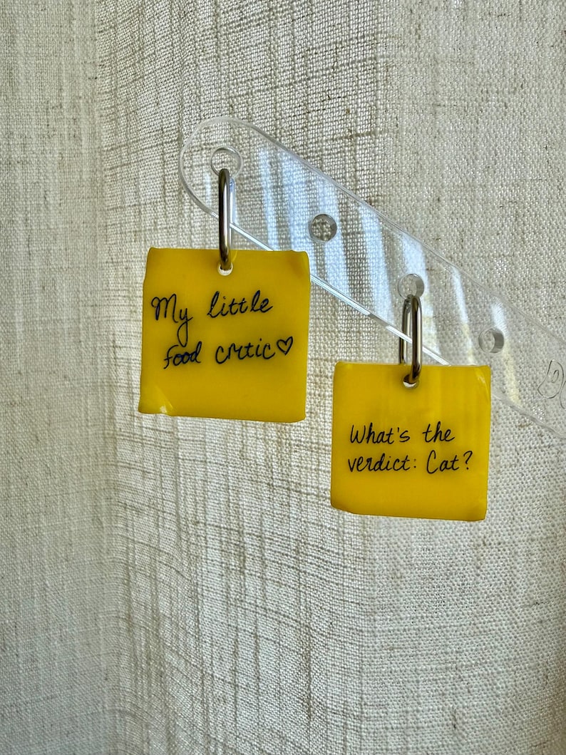 Op de afbeelding: Een paar gele vierkante oorbellen met zilveren ringen. De ene oorbel bevat de zwarte tekst "My little food critic" met een hartje, en de andere oorbel bevat de zwarte tekst "What's the verdict: Cat?" in een handgeschreven stijl.