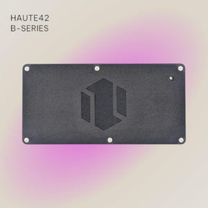 May include: A black rectangular device with the text "HAUTE42 B-SERIES" at the top. The device has a recessed geometric design in the center and is secured with silver screws at each corner. The background has a gradient of white to pink.