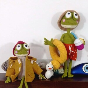 May include: Two crocheted frog plushies, one wearing a yellow raincoat and the other wearing a red and white swimsuit and holding a yellow inflatable ring. A white crocheted chick sits in front of the frogs.