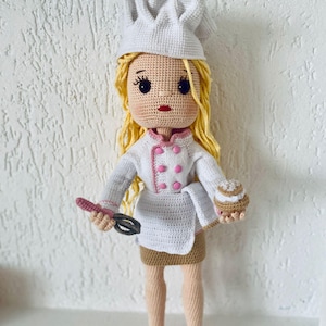 May include: A crocheted doll wearing a white chef's hat and a white and pink chef's uniform. The doll is holding a whisk and a small cake.