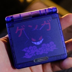 Könnte beinhalten: Ein lila Nintendo Game Boy Advance SP Handheld-Konsole mit einem Gengar-Design. Das Design umfasst japanischen Text und eine lächelnde Gengar-Illustration in Lila. Die Konsole hat ein lila Scharnier und ein silbernes Nintendo-Logo.