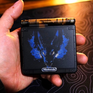 Puede incluir: Una consola Nintendo Game Boy Advance SP negra con un diseño de lobo azul y negro en la parte delantera. El logotipo de Nintendo es visible en la parte inferior del dispositivo.