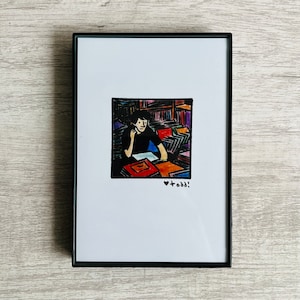 May include: A framed illustration of a person sitting at a desk surrounded by colorful books. The person is wearing a black shirt and has dark hair. The illustration is in a black frame and has the text "todd!" written in black below the image.