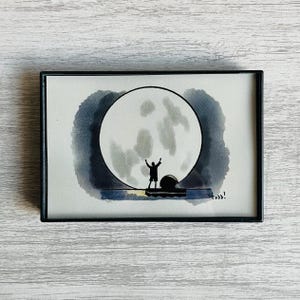 May include: A framed watercolor illustration featuring a black silhouette of a person standing with arms raised against a large, pale gray full moon. The scene includes a dark blue horizon line and the handwritten signature "todd!" in the corner.