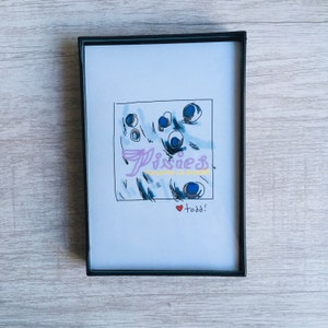 May include: A framed print with a blue and white abstract design. The print has the text "Pixies" and "Across the Universe" in pink and yellow. The print also has the text "todd!" with a red heart.