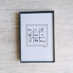 May include: Framed black and white artwork featuring a hand-drawn tic-tac-toe board with the words "TRIO", "AND", and "ERROR". The board includes various symbols and the artist's signature.