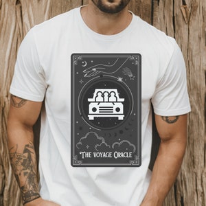 Puede incluir: Camiseta blanca con un gráfico en blanco y negro de una carta de tarot. La carta presenta un coche con tres personas dentro, una mano que se extiende desde el cielo y el texto "The Voyage Oracle".