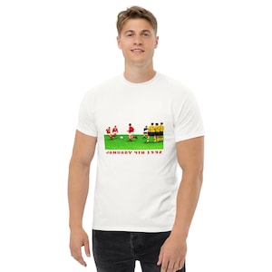 May include: A white short-sleeved t-shirt featuring a graphic of soccer players on a green field with the text JANUARY 4TH 1992 printed in red below the image. The graphic depicts players in red and yellow uniforms during a match.