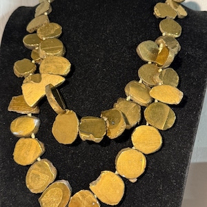 May include: A two-strand necklace featuring irregularly shaped, gold-colored beads. The beads are strung together with a neutral-colored cord. The necklace is displayed on a black mannequin.