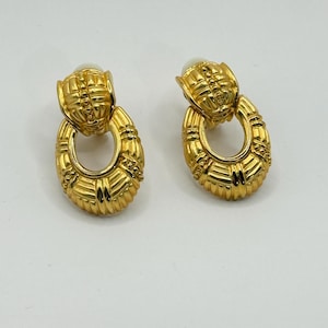 以下が含まれることがあります： 金色のイヤリングのペア。各イヤリングは、テクスチャ加工された楕円形のドロップと、より小さなテクスチャ加工された上部を備えています。各イヤリングの上部にはイミテーションパールが付いています。イヤリングはヴィンテージ感があります。