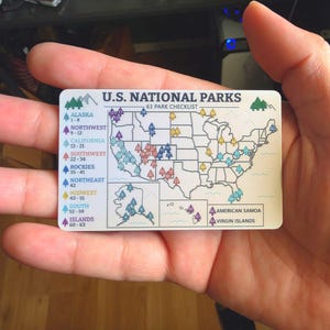 May include: A rectangular white card featuring a U.S. National Parks 63 park checklist map. The map displays the United States with numbered tree icons color-coded by region, including Alaska, Hawaii, and territories, with a legend on the left.