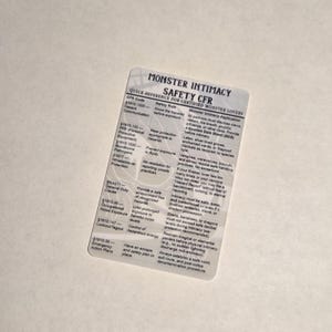 May include: A clear plastic card with the text "MONSTER INTIMACY SAFETY CFR" in black. The card provides a quick reference for certified monster lovers, listing safety rules and CFR codes. The card is on a white surface.
