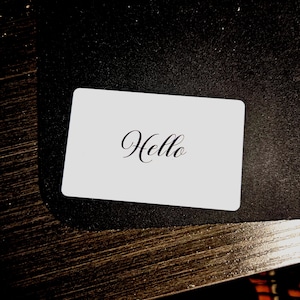 May include: A white rectangular card with the word "Hello" written in elegant cursive script. The card is set against a dark, textured background, creating a contrast that highlights the text.