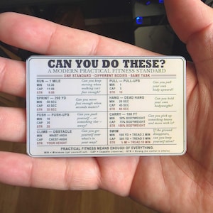 May include: A rectangular white fitness reference card with black text titled CAN YOU DO THESE? A MODERN PRACTICAL FITNESS STANDARD. The card lists various physical tasks like running, sprinting, push-ups, climbing, pull-ups, hanging, carrying, and swimming.