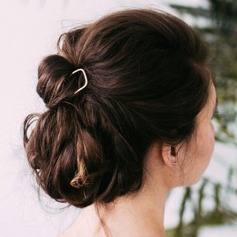 May include: A woman's dark brown hair styled in a low bun, secured with a gold-colored hair pin. The hair pin is a simple, curved design. The background is a soft, neutral color.