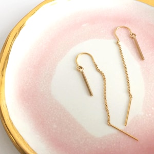 May include: A pair of gold threader earrings. Each earring has a straight bar attached to a fine chain that passes through the ear. The earrings are presented on a pink and white plate with a gold rim.