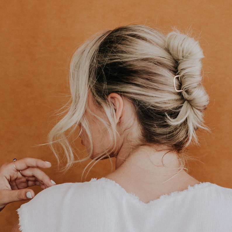 May include: A gold-colored square hair clip secures a blonde bun hairstyle. The hair is styled in a loose, textured updo. The background is a solid, warm orange-brown color.