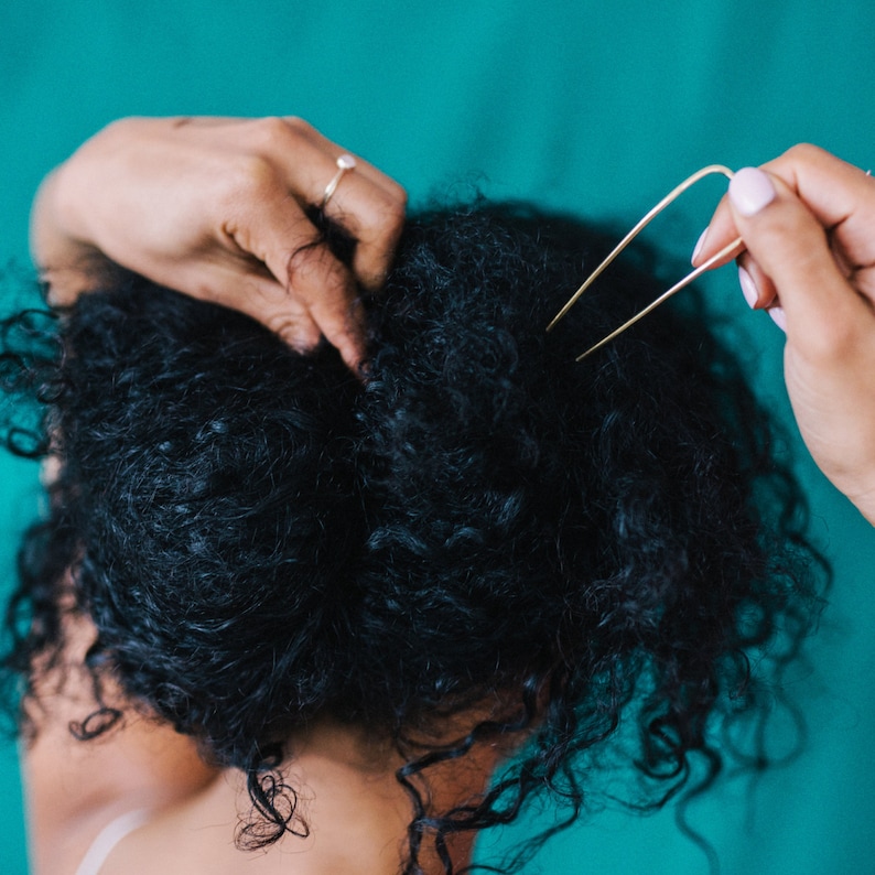 May include: A person's dark, curly hair is styled with a gold hair pin. The person's hand with a ring is holding the hair, while another hand holds the pin. The background is a solid teal color.
