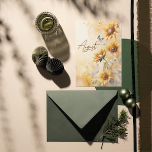 May include: A green envelope with a card featuring a watercolor painting of sunflowers and the word "August". The card is surrounded by decorative ornaments and a sprig of greenery.