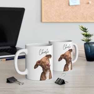 Op de afbeelding: Twee witte keramische mokken met een bruin hondenhoofd op elk. De mok aan de linkerkant heeft de naam "Charlie" erop gedrukt. De mok aan de rechterkant heeft de naam "Charlie" erop gedruckt.
