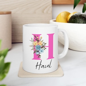 Peut inclure: Mug en céramique blanche avec une lettre H rose et un motif floral. Le nom Hazel est écrit sous la lettre.