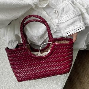 Può includere: Una borsa tote bordeaux intrecciata con manici intrecciati. La borsa presenta un motivo a trama di cestino e una forma strutturata. L'interno sembra avere una fodera di colore chiaro. La borsa è un accessorio elegante.