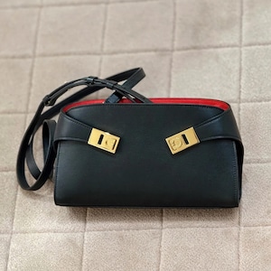 May include: Black rectangular handbag with a red interior and gold-colored hardware. The bag has a long, adjustable strap and two gold clasps on the front. The bag is made of a smooth material.