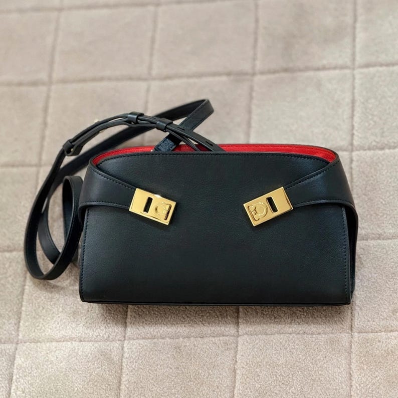 May include: Black rectangular handbag with a red interior and gold-colored hardware. The bag has a long, adjustable strap and two gold clasps on the front. The bag is made of a smooth material.