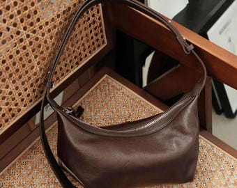 May include: A dark brown hobo bag with a smooth, slightly textured surface, a curved top zipper closure with a silver-toned metal pull, and an adjustable shoulder strap with a metal buckle, featuring a rounded, slouchy silhouette.