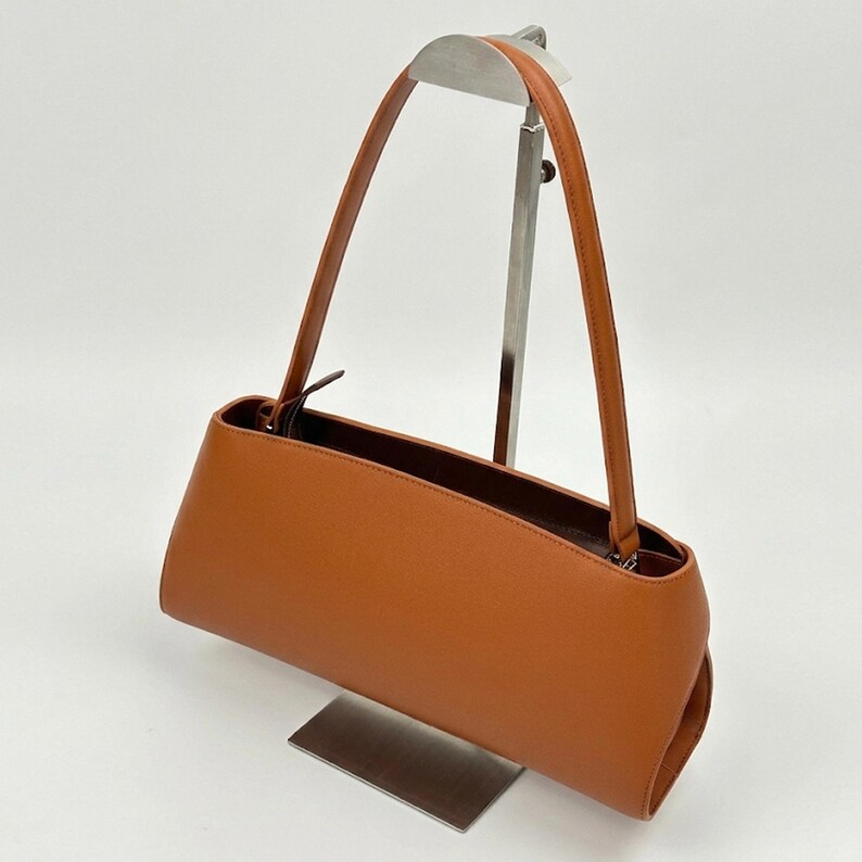 May include: A rectangular, tan-colored handbag with a long, thin strap. The bag is made of a smooth material and has a simple, elegant design. The bag is displayed on a metal stand against a white background.