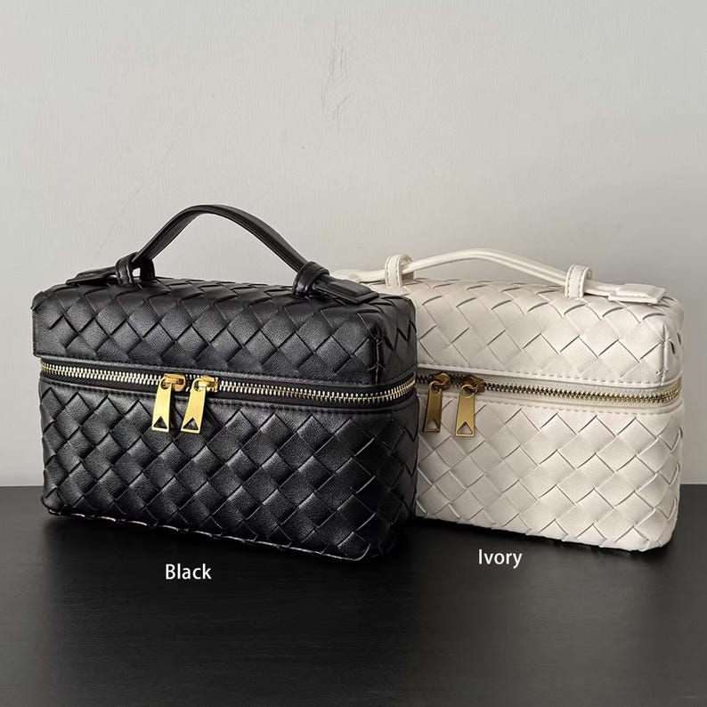 Puede incluir: Se muestran dos neceseres de cuero tejido, uno negro y otro marfil. Cada bolso tiene forma rectangular, un asa superior y una cremallera dorada. La bolsa negra tiene la palabra "Black" debajo y la bolsa marfil tiene la palabra "Ivory" debajo.