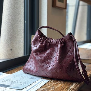 May include: A burgundy leather handbag with a woven design. The bag features a drawstring closure and a single handle. It rests on a wooden surface near a window. The bag's colour is a deep red, almost maroon.