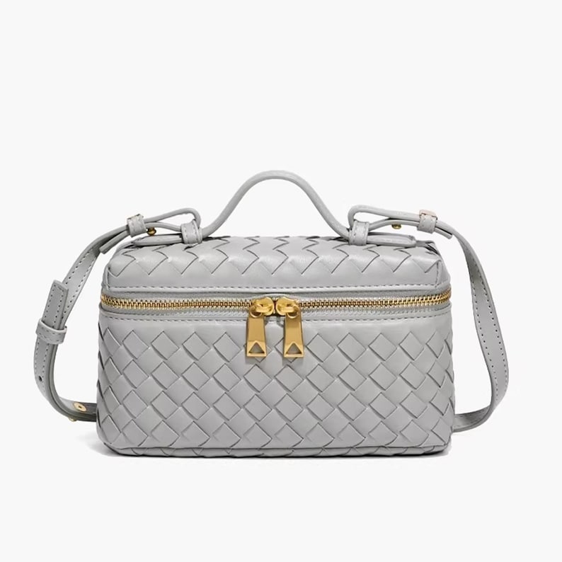 Puede incluir: Un bolso bandolera de cuero tejido gris claro con un asa superior y una cremallera dorada. El bolso tiene forma rectangular y una correa para el hombro desmontable. El bolso est&aacute; dise&ntilde;ado con un patr&oacute;n de tejido de cesta.