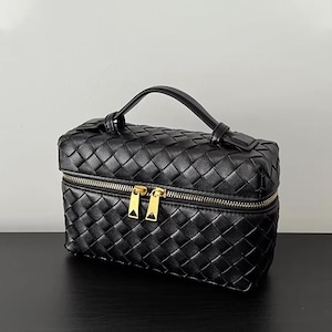 Puede incluir: Bolso de cosm&eacute;ticos de cuero negro tejido con asa superior y cremallera dorada. El bolso tiene forma rectangular y un patr&oacute;n tejido. La cremallera tiene dos tiradores. El bolso est&aacute; sobre una superficie negra.