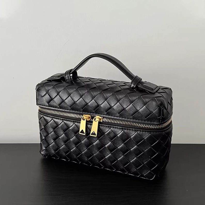 Puede incluir: Bolso de cosm&eacute;ticos de cuero negro tejido con asa superior y cremallera dorada. El bolso tiene forma rectangular y un patr&oacute;n tejido. La cremallera tiene dos tiradores. El bolso est&aacute; sobre una superficie negra.