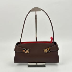 May include: A brown leather handbag with a red interior. The bag features a rectangular shape with a flap closure and gold-tone hardware. The handles are thin and arched. The bag is displayed on a silver stand.