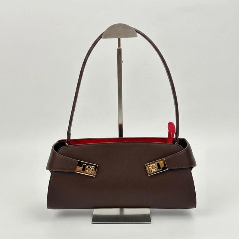 May include: A brown leather handbag with a red interior. The bag features a rectangular shape with a flap closure and gold-tone hardware. The handles are thin and arched. The bag is displayed on a silver stand.