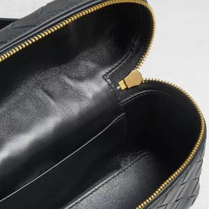 Puede incluir: Primer plano de un bolso de mano de cuero negro con cremallera dorada. El interior es visible, revelando una superficie oscura y lisa. La cremallera est&aacute; parcialmente abierta, mostrando el contenido del bolso.