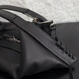 May include: A black handbag featuring a smooth matte finish, structured top handles, and a silver-toned metal zip closure. The side panel displays a decorative braided leather detail, and a small silver metal D-ring is attached to the upper corner.