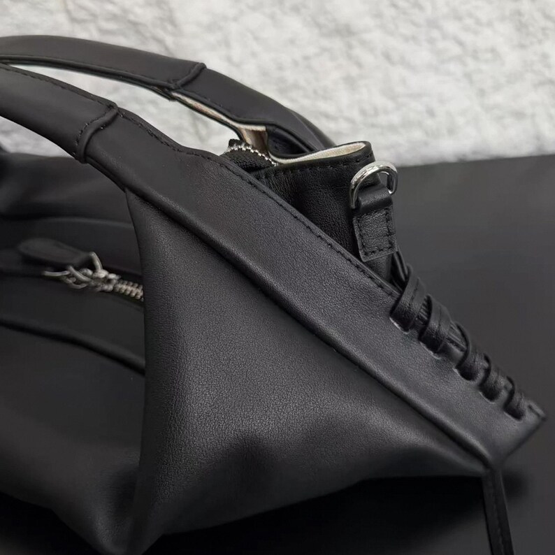 May include: A black handbag featuring a smooth matte finish, structured top handles, and a silver-toned metal zip closure. The side panel displays a decorative braided leather detail, and a small silver metal D-ring is attached to the upper corner.