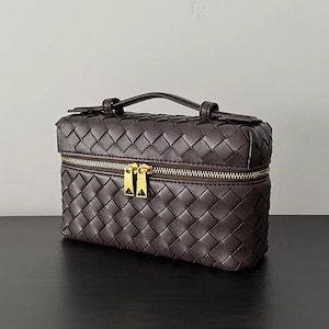 Puede incluir: Un neceser de cuero tejido marr&oacute;n oscuro con un asa superior y una cremallera dorada. La bolsa es rectangular y presenta un patr&oacute;n tejido repetido. La cremallera tiene dos tiradores. La bolsa est&aacute; sobre una superficie negra.