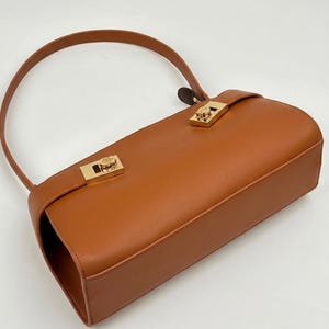 May include: A tan leather handbag with a curved handle and gold-colored hardware. The rectangular bag features a flap closure with a decorative clasp. The bag's design is simple and elegant, suitable for everyday use.