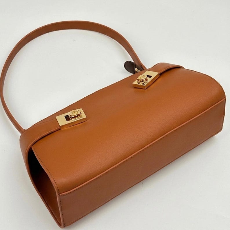 May include: A tan leather handbag with a curved handle and gold-colored hardware. The rectangular bag features a flap closure with a decorative clasp. The bag's design is simple and elegant, suitable for everyday use.