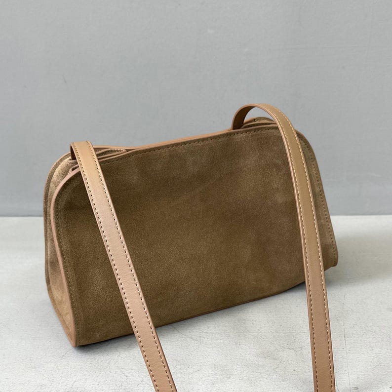 May include: A rectangular, brown suede handbag with a long, beige shoulder strap. The bag has a structured shape with rounded corners and a contrasting beige trim. The strap is thin and appears to be made of leather.
